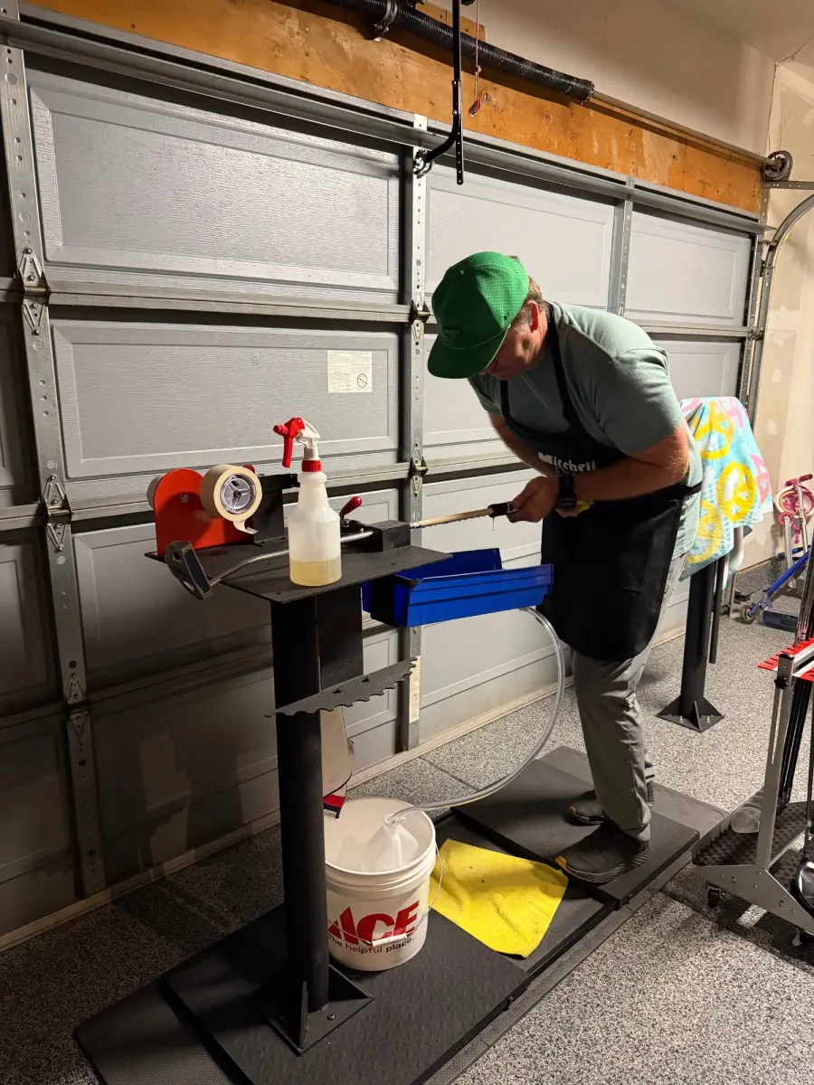 Kyle at his regripping station in the garage, working a new grip onto a club over a blue solvent tray, with a spray bottle and bucket beside him